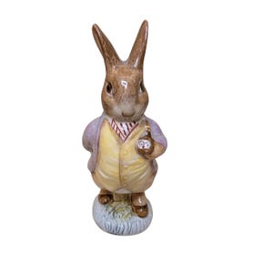 May include: A ceramic figurine of a brown rabbit standing upright on a circular base. The rabbit wears a light purple jacket over a yellow vest with a red and white striped necktie. One paw is raised, holding a small brown object with floral details.