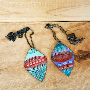 May include: Two leather pendants on chains, one is teal with red and white accents, the other is purple with red and white accents.