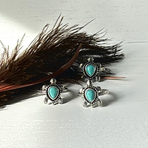 May include: Three silver-toned rings with turquoise turtle designs. Each ring features a turtle-shaped setting with a teardrop-shaped turquoise stone. The rings are displayed on a white surface with a brown feather.