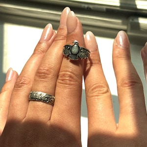 Könnte beinhalten: Ein silberner Ring mit einem Vogeldesign und einem kleinen ovalen Stein wird an einem Finger getragen. Ein weiterer silberner Ring mit einem floralen Design ist ebenfalls sichtbar. Ein dünner silberner Bandring befindet sich an einem anderen Finger. Die Ringe werden auf einer Hand präsentiert.