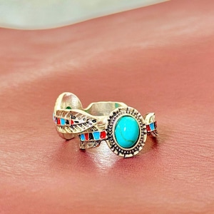 May include: Silver-toned ring featuring a large, oval turquoise stone at the center. The ring has decorative feather-like details with red, blue, and black accents. The ring is set against a pink background.