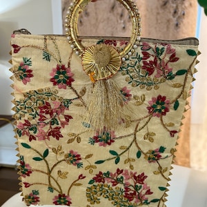 May include: A small, cream-colored clutch bag with a floral pattern. The bag has a gold ring handle with white pearls and a gold tassel.