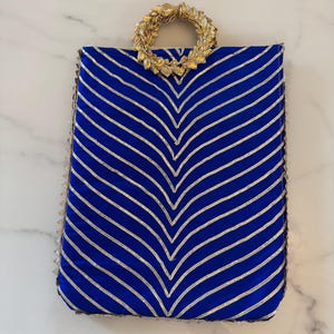 May include: A royal blue handbag with a gold-colored handle and trim. The bag features a chevron pattern created by gold-colored stripes. The handle is a circular design made of gold-colored leaves.