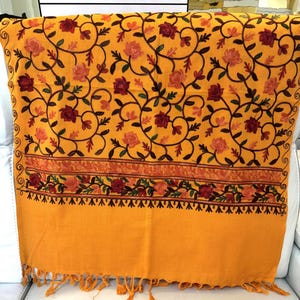 May include: A vibrant mustard yellow shawl featuring intricate floral embroidery in shades of deep red, orange, and dark green. The design includes swirling vine patterns across the main body and a horizontal border with fringe at the bottom.
