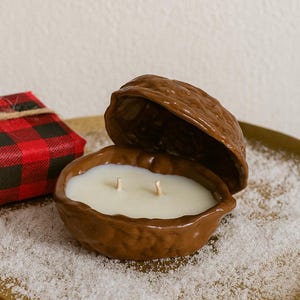 Puede incluir: Una vela de cerámica marrón con forma de nuez, con una vela blanca y dos mechas. La parte superior de la nuez está abierta. Un pequeño regalo envuelto en papel a cuadros rojo y negro está a la izquierda. La vela está sobre una bandeja dorada.