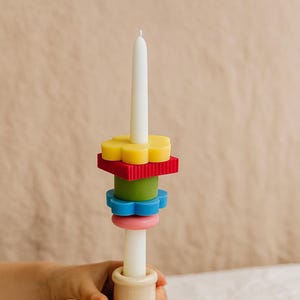 Puede incluir: Una vela blanca está centrada en un candelabro beige, adornada con formas de cera coloridas. Las formas incluyen una flor amarilla, un cuadrado rojo, una flor verde, una flor azul y un círculo rosa. También hay dos piezas de cera decorativas adicionales.