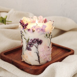 May include: A white candle with dried flowers embedded in the wax. The candle is lit and the flame is visible. The candle is sitting on a wooden tray on a white blanket.