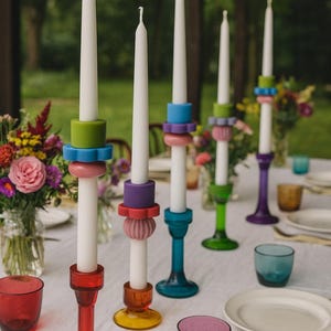 Puede incluir: Una colección de coloridos candelabros de vidrio con velas blancas. Los candelabros son de varios colores, incluyendo rojo, naranja, verde, azul y morado. La mesa está puesta con vasos y platos.
