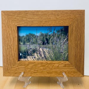 May include: A framed photograph featuring a field of lavender under a bright blue sky. The image is enclosed in a rectangular, light brown wooden frame. The frame is supported by two clear plastic stands.