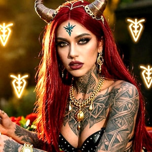 May include: A woman with vibrant red hair, styled with gold horns and a headpiece. She is wearing a black top and gold jewellery, including a necklace and earrings. Her skin is tattooed, and she has dark makeup.
