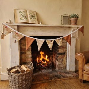 May include: A fabric bunting banner featuring alternating triangular flags in solid burnt orange, cream and brown striped patterns, and a vintage-style botanical print on a white string, displayed across a white wooden fireplace mantel.