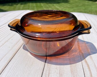 May include: Amber-colored glass casserole dish with a matching lid. The dish has two handles and is sitting on a white wooden surface. The lid has a textured design. The background is a green lawn and trees.