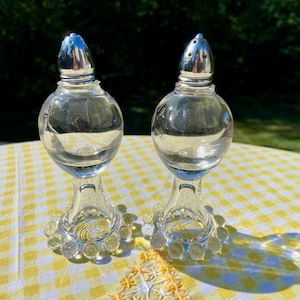 May include: A pair of clear glass salt and pepper shakers. Each shaker has a round body, a silver metal top, and a decorative base with small glass circles. The shakers are on a yellow and white checkered tablecloth.