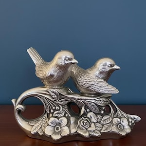 May include: A silver-colored decorative sculpture featuring two birds perched on a floral branch. The birds have detailed feather textures and are set against a backdrop of flowers and leaves. The sculpture is made of metal.