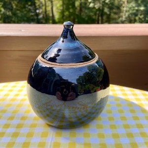 May include: A handmade ceramic jar with a lid. The jar is a dark blue color with a glossy finish, and the bottom half has a lighter, textured pattern. The jar is sitting on a yellow and white checkered tablecloth.