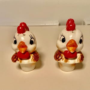 May include: A pair of ceramic salt and pepper shakers shaped like cartoon chickens. The chickens have white bodies, red combs and wattles, orange beaks, and brown and white feathered details. The shakers are designed to sit upright.