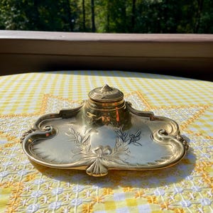 May include: A vintage brass inkwell with a decorative floral design. The inkwell has a lid and sits on a tray with ornate edges. The inkwell is displayed on a yellow and white checkered tablecloth, with a blurred green background.