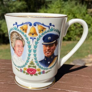 May include: White ceramic mug with a gold rim and handle. The mug features portraits of a man and woman, with gold bells and floral accents. The mug is likely a commemorative item.