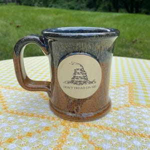 May include: A ceramic mug with a speckled brown and dark blue glaze featuring a circular beige decal on the front. The decal displays a black line drawing of a coiled rattlesnake above the text DON'T TREAD ON ME in a simple sans-serif font.