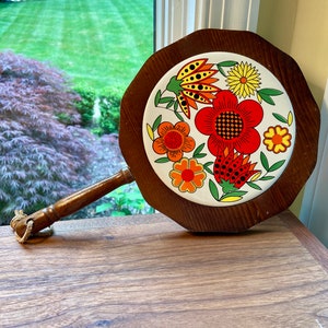May include: A wooden serving tray with a round, white ceramic tile inset. The tile features a colorful floral design with red, orange, yellow, and green flowers. The tray has a wooden handle.