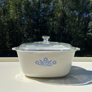 May include: A white casserole dish with a clear glass lid. The dish features a blue floral design on the front. The lid has a clear glass knob. The dish is approximately 9 inches wide.