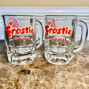 May include: Two clear glass mugs with handles. Each mug has the word "Frostie" in red script with a cartoon character and the words "Root Beer" in red block letters. The mugs are on a brown and tan marble-patterned surface.
