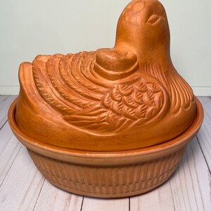 May include: A terracotta-colored duck-shaped casserole dish. The lid features detailed feather carvings and a duck head. The base has a ribbed design. This decorative dish is suitable for serving or display.
