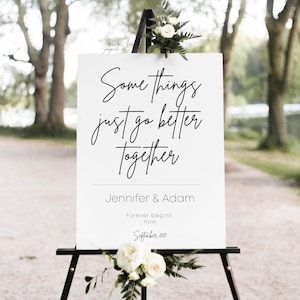 Puede incluir: Un cartel de boda blanco con caligrafía negra que dice "Some things just go better together." Debajo de la caligrafía está el texto "Jennifer & Adam" y "Forever begins now" con la fecha "September 2023".