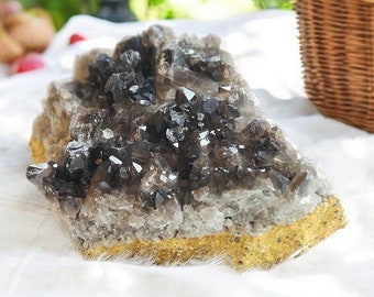 May include: A large, smoky quartz crystal cluster with many points. The crystal is a light brown color with a clear, glassy finish. The crystal is sitting on a white cloth.