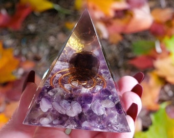 May include: A clear glass pyramid filled with amethyst crystals and a copper wire spiral. The pyramid is sitting on a hand.