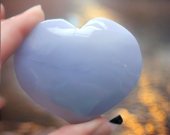 May include: A heart-shaped blue chalcedony crystal, polished smooth and shiny. The crystal is held in a hand, with the background blurred.