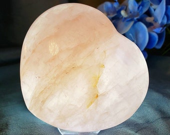 May include: A large, heart-shaped, polished rose quartz crystal with a light pink color and white streaks. The crystal is sitting on a clear stand.