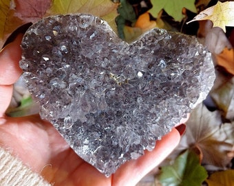 May include: A heart-shaped crystal cluster with a clear, glassy appearance. The crystal is made up of many small, pointed crystals that are clustered together. The crystal is a light gray color with a hint of purple.