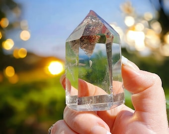 May include: A clear quartz crystal point with a natural inclusion. The crystal is held in a hand against a blurred background of blue sky and green foliage.