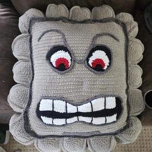 May include: A gray crocheted pillow with a grumpy face. The face has black eyebrows, red and white eyes, and a white and black mouth.