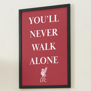 Könnte beinhalten: Gerahmtes Wandbild mit rotem Hintergrund und den weißen Worten "YOU'LL NEVER WALK ALONE". Das Emblem des Liverpool Football Clubs befindet sich unten. Der Rahmen ist schwarz.