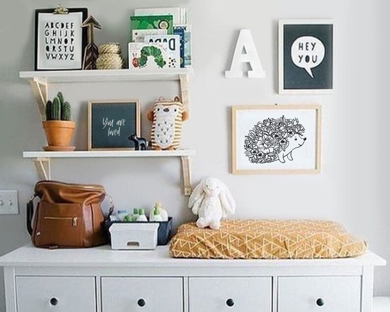 May include: A white dresser with four drawers, a changing pad with a yellow and brown geometric pattern, and a white shelf with various items, including a black and white print of a hedgehog with flowers, a black chalkboard with the text "You are loved", a brown leather diaper bag, and a white stuffed bunny.