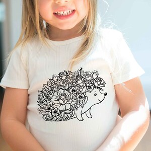 May include: A white t-shirt with a black and white illustration of a hedgehog with flowers on its back.