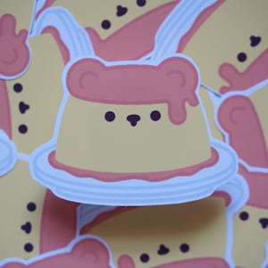 May include: A cute cartoon sticker of a yellow pudding with a pink bear face and pink dripping sauce on top. The pudding is on a white plate.