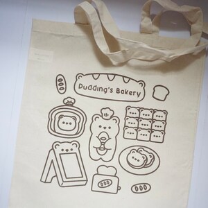 May include: A white canvas tote bag with brown printed designs. The bag features a bear wearing a chef hat holding a loaf of bread, a sign that says "Pudding's Bakery", and other bakery-themed illustrations.