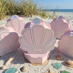 May include: Pink seashell-shaped boxes with iridescent accents, arranged on a sandy beach with seashells, sea glass, and a starfish. The boxes have a pearl-like embellishment on top, evoking a coastal theme.