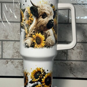 May include: White insulated tumbler with a handle, featuring a watercolor design of a Highland cow surrounded by sunflowers. The tumbler has a clear lid and is approximately 25 cm tall.