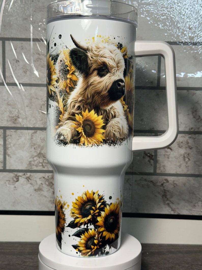 May include: White insulated tumbler with a handle, featuring a watercolor design of a Highland cow surrounded by sunflowers. The tumbler has a clear lid and is approximately 25 cm tall.