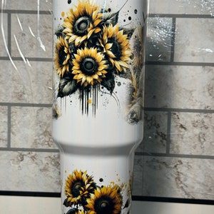 May include: A white insulated tumbler with a sunflower design. The sunflowers are yellow and black, with green leaves and black paint splatter. The tumbler has a clear lid. The design wraps around the tumbler.
