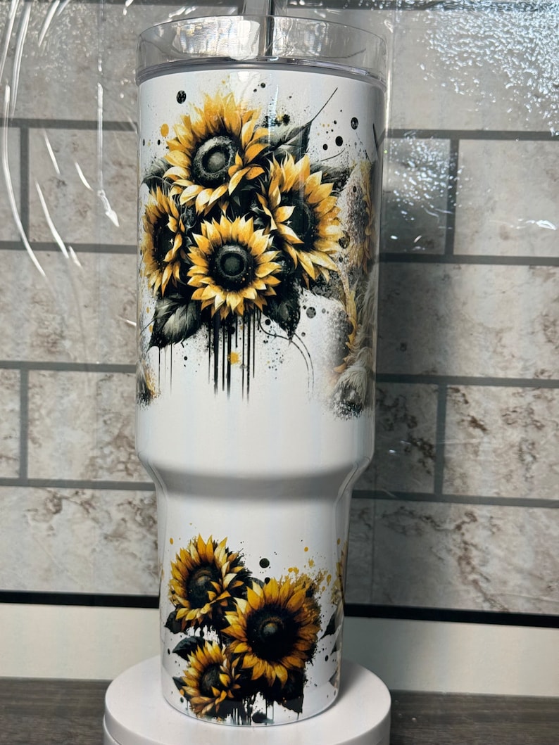 May include: A white insulated tumbler with a sunflower design. The sunflowers are yellow and black, with green leaves and black paint splatter. The tumbler has a clear lid. The design wraps around the tumbler.