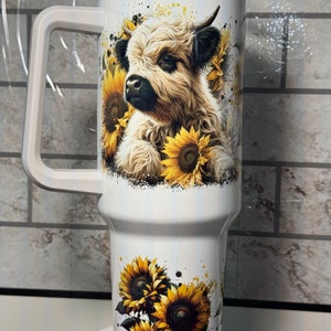 May include: White insulated travel mug with a handle, decorated with a Highland cow and sunflowers. The design features yellow sunflowers and a watercolour effect. The mug has a clear lid and is designed for drinks.
