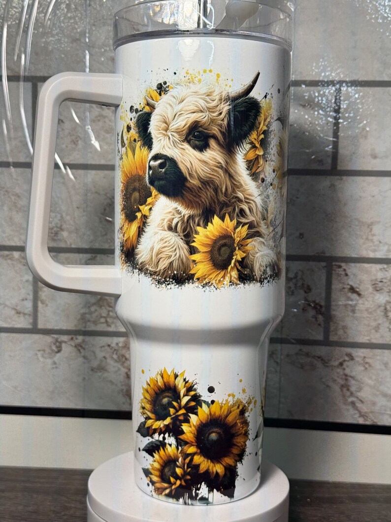 May include: White insulated travel mug with a handle, decorated with a Highland cow and sunflowers. The design features yellow sunflowers and a watercolour effect. The mug has a clear lid and is designed for drinks.