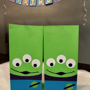 May include: Two green gift bags featuring the Little Green Men from Toy Story. Each bag has three white eyes, a black smile, and a blue and purple section at the bottom with a green and black design. A birthday banner is in the background.