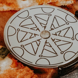 May include: A close-up of a pizza cutter with a black handle and a silver wheel with the word "pizza" repeated around the edge. The cutter is on a wooden chopping board with a slice of pizza.