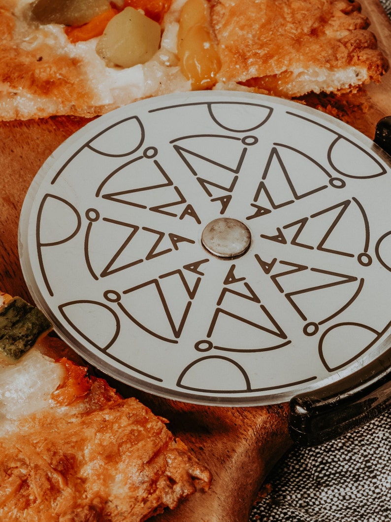 May include: A close-up of a pizza cutter with a black handle and a silver wheel with the word "pizza" repeated around the edge. The cutter is on a wooden chopping board with a slice of pizza.